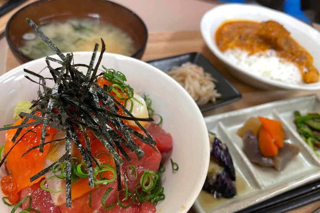 色とりどりの新鮮な海鮮がたっぷりのった海鮮丼を中心に、味噌汁、小鉢、スパイシーチキンカレーなどが並ぶ豪華な定食メニュー。府中市場内の「のんしゃらん食堂」で提供されるランチセットの一例で、刺身、いくら、刻みネギ、海苔が美しく盛り付けられている。