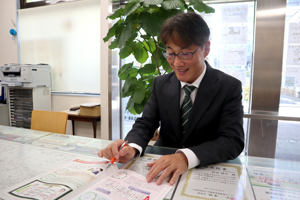 スーツ姿の男性がガラス張りの明るいオフィスで冊子を広げながらペンを手に持ち、楽しそうに説明している様子。テーブルには認定証や案内資料が並び、背景には観葉植物とプリンターが置かれた落ち着いた室内空間。