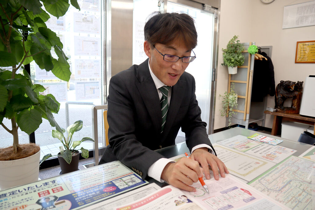 スーツ姿の男性が明るい不動産オフィスでパンフレットを見ながら説明する様子。テーブルには地図や資料が並び、背景には観葉植物やインテリアが配置されている。