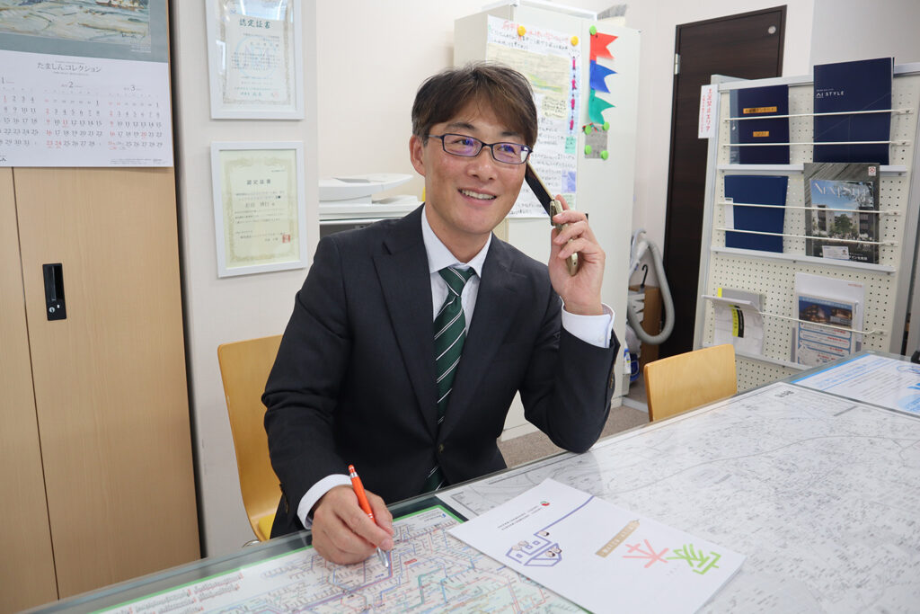 スーツ姿の不動産営業マンがオフィスで電話をしながら地図を確認している様子。デスクには物件資料や路線図が広がり、背後には証明書やパンフレット棚が並んでいる。