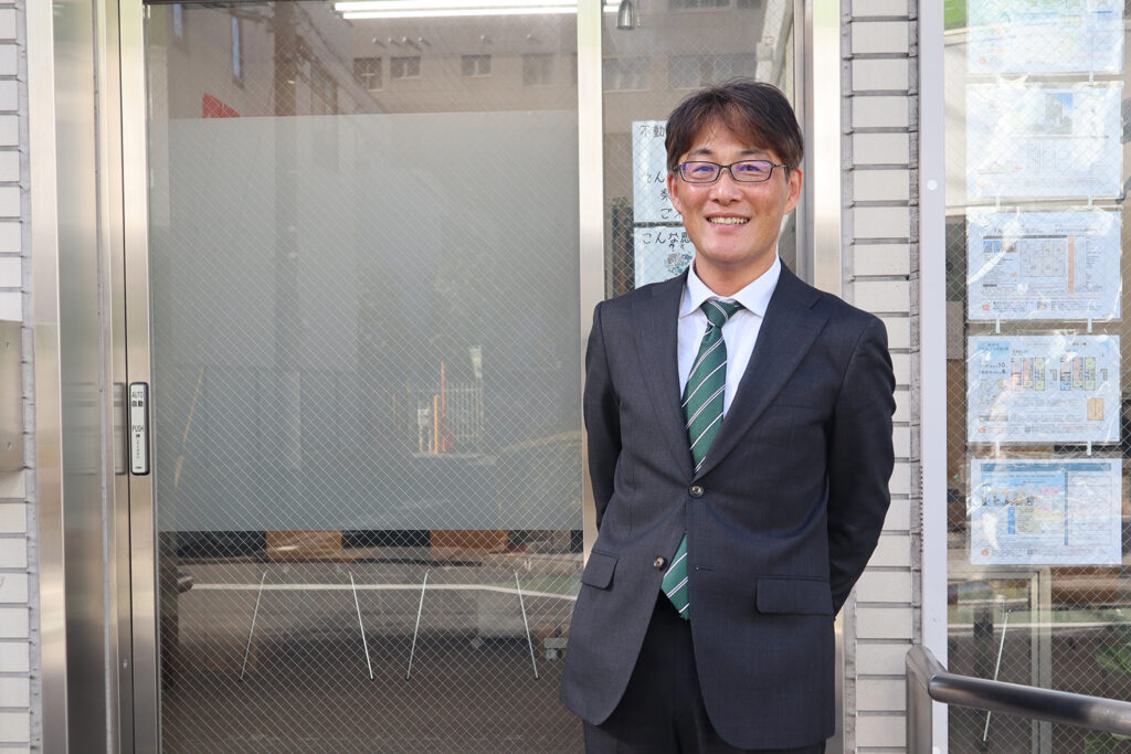 スーツ姿の不動産営業マンが店舗前で笑顔を見せるポートレート写真。店舗の窓には物件情報が掲示されており、信頼感のある雰囲気が伝わる。