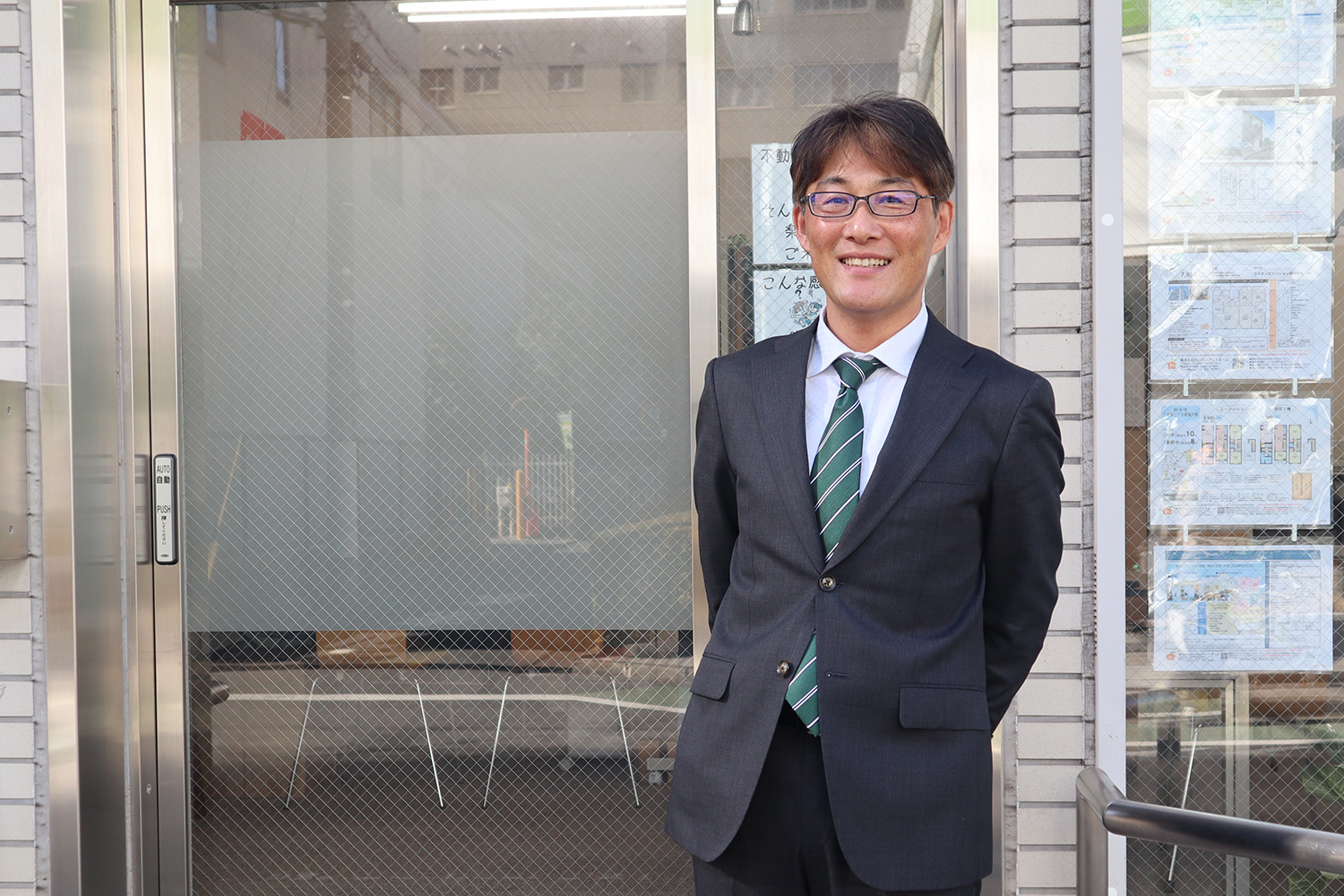 スーツ姿の不動産営業マンが店舗前で笑顔を見せるポートレート写真。店舗の窓には物件情報が掲示されており、信頼感のある雰囲気が伝わる。