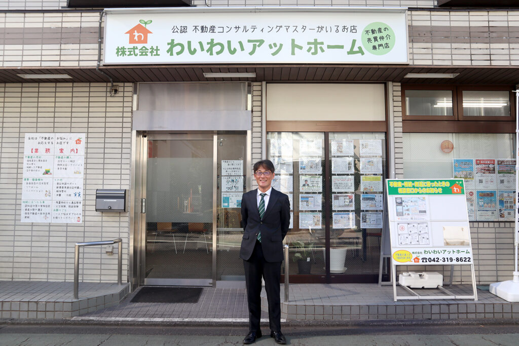 「株式会社わいわいアットホーム」の店舗前に立つスーツ姿の男性スタッフ。不動産売買・仲介を専門とする公認不動産コンサルティングマスターのいる信頼のお店。