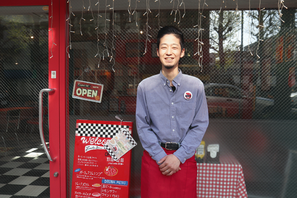赤い扉の前で笑顔の男性スタッフが立っているレストランの外観。ガラスには「OPEN」のサインと営業時間・メニューが書かれた赤い看板があり、チェック柄のテーブルクロスも見える。店内は黒と白の市松模様の床で、アメリカンダイナー風の雰囲気。