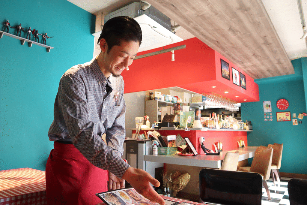 アメリカンダイナー風の店内で、赤いエプロンを着けた男性スタッフがメニューを指差しながら案内している様子。背景には赤とターコイズブルーの壁、バーカウンター、天井に吊るされたグラス、棚に並んだフィギュアが見える。