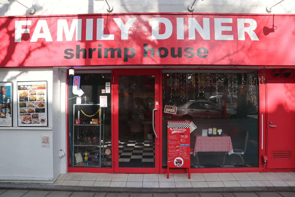 赤い外観が目を引くレストラン「FAMILY DINER shrimp house」の正面。ガラス扉には「OPEN」のサインがあり、店内の市松模様の床と赤いギンガムチェックのテーブルクロスが見える。外壁にはメニュー写真が掲示されている。