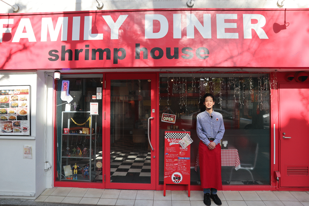 赤い外観のレストラン「FAMILY DINER shrimp house」の正面で、赤いエプロンを着けた男性スタッフが立っている様子。ガラス扉には「OPEN」のサインとメニュー看板があり、店内の市松模様の床と赤いギンガムチェックのテーブルクロスが見える。