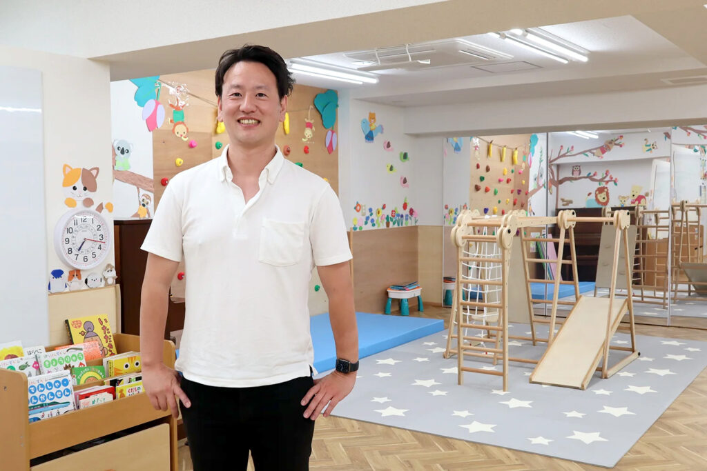 室内遊具や滑り台、カラフルなボルダリング壁がある保育施設の中で、白いシャツの男性が笑顔で立っている。背後には本棚や時計、動物の壁面装飾がある。