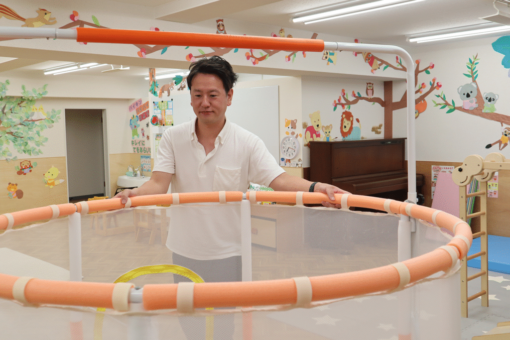 保育園や幼稚園の室内で、白いシャツの男性がオレンジ色の縁取りがあるネット付き遊具を両手で支えている。周囲には動物キャラクターやピアノが配置された明るい室内。