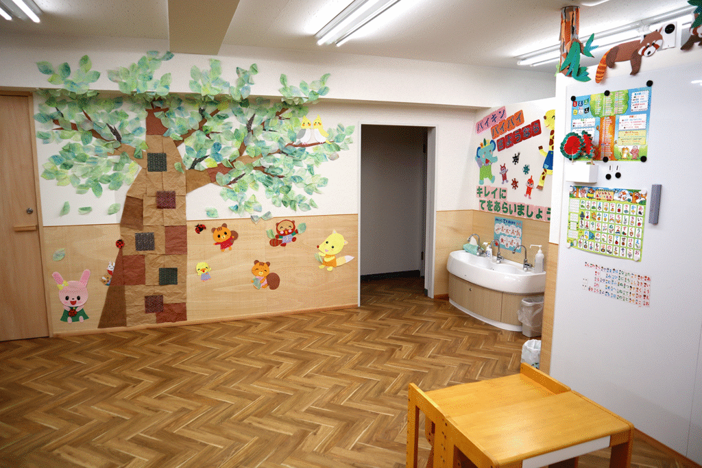 幼児施設の一角。大きな木の壁面装飾と動物キャラクター、子供用の洗面台が配置されている。壁には「バイキン バイバイ きれいにてをあらいましょう」と書かれたポスターが貼られている。