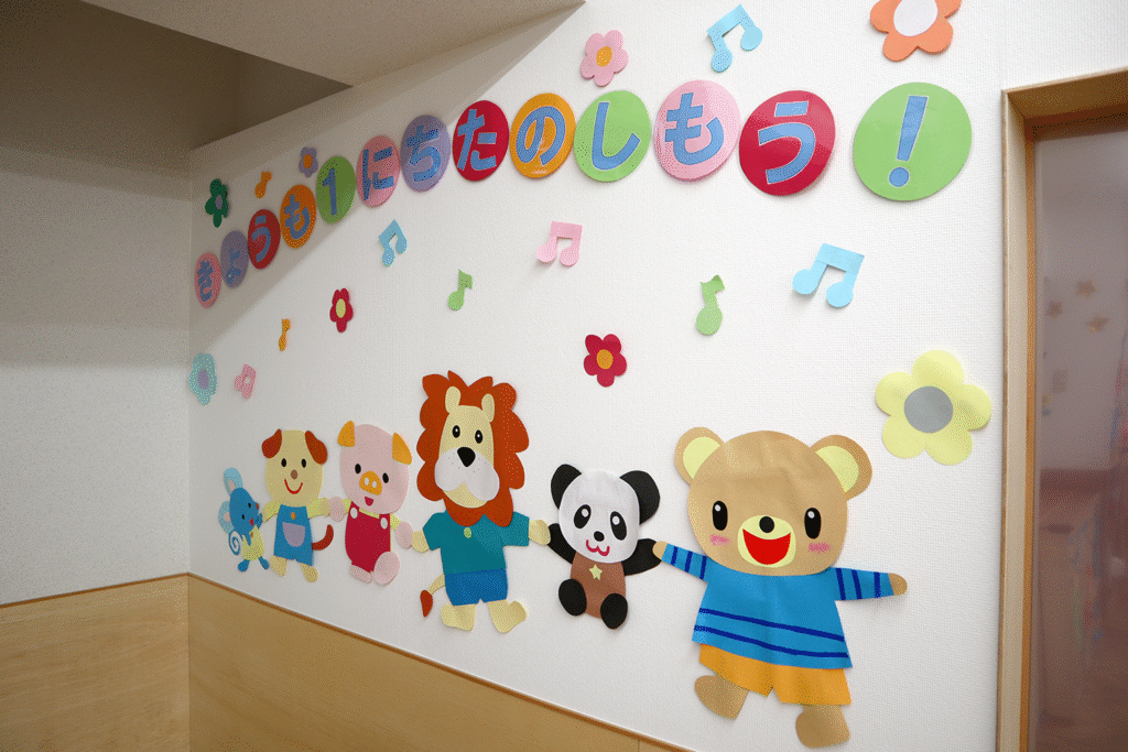 壁に貼られたカラフルな動物キャラクターと「きょうも1にちたのしもう！」と書かれたメッセージ。花や音符の装飾が散りばめられている幼稚園や保育園の一角。