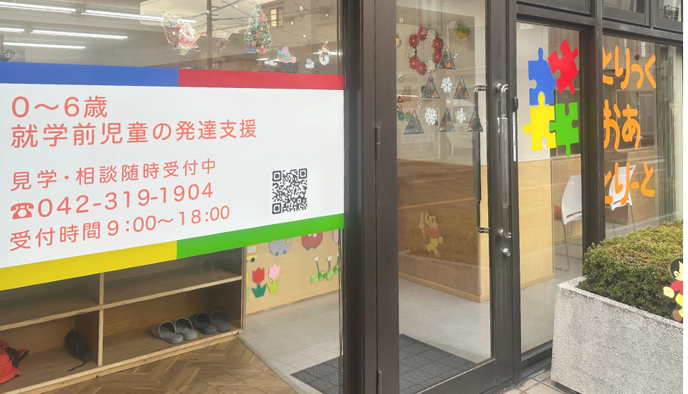 ガラス張りの入口がある幼児向け施設の外観。窓には「0～6歳 就学前児童の発達支援」「見学・相談随時受付中」「受付時間9:00～18:00」などの案内と電話番号が書かれた看板が貼られている。入口横のガラスにはカラフルなパズルピースの装飾と文字が見える。室内には季節の飾りや子供向けの装飾が施されている。