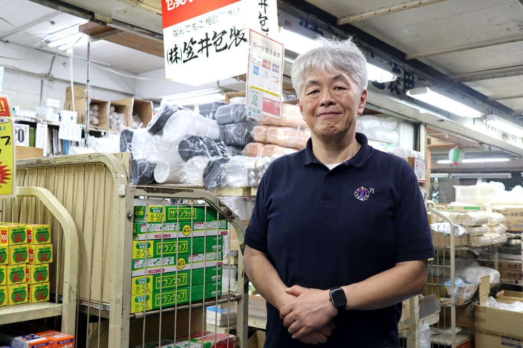 包装資材が並ぶ店内で、紺色のポロシャツを着た白髪の男性が笑顔で立っている。背後にはラップや容器などの商品が陳列されており、上部には「株式会社笠井包装」と書かれた看板が見える。