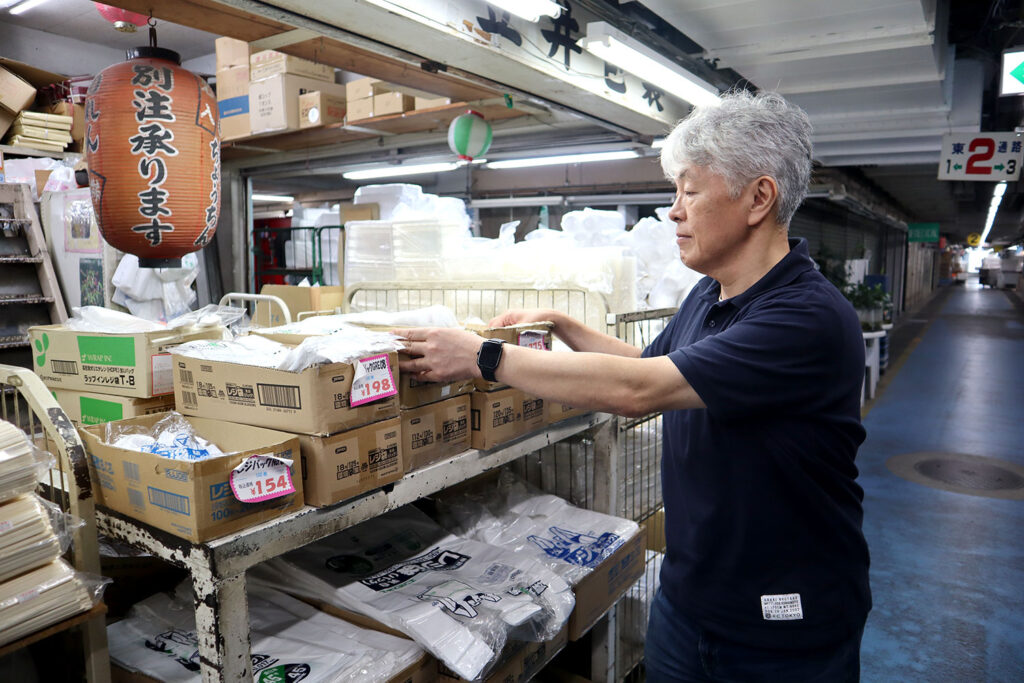 紺色のポロシャツを着た白髪の男性が、包装資材が入った段ボール箱を棚に整理している。店内には「別注承ります」と書かれた提灯が吊るされ、周囲にはビニール袋や箱詰めの商品が並んでいる。奥には長い通路と照明が続く市場のような雰囲気がある。