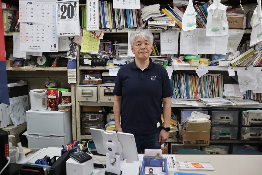 オフィスのカウンター前で、紺色のポロシャツを着た白髪の男性が正面を向いて立っている。背景には本や書類がぎっしりと詰まった棚があり、カレンダーやメモが壁に貼られている。机の上にはレジ端末や書類、小物が置かれており、実務的な雰囲気が漂っている。