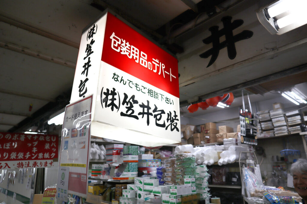 店内の天井から吊るされた「包装用品のデパート 株式会社笠井包装」の看板が明るく照らされている。看板の下には「なんでもご相談下さい」と書かれており、近くにはクレジットカードや電子マネー対応を示す案内も掲示されている。背景には梱包資材や段ボール箱、ラップなどの商品が所狭しと並んでいる。