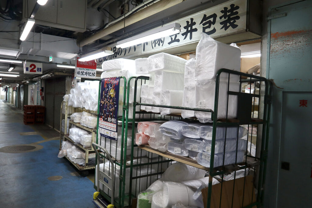 「株式会社笠井包装」の店舗前に、発泡スチロール容器や包装資材が積まれた台車が並んでいる。看板には「包装用品のデパート」と書かれており、通路には「東2通路」の標識も見える。店内には白い梱包材や段ボールが所狭しと保管されている様子がうかがえる。