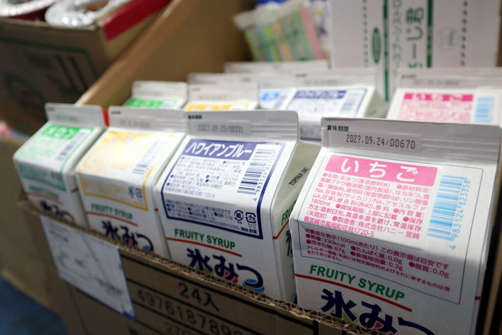 段ボール箱の中に並べられた複数のフルーツシロップの紙パック。手前には「いちご」味と「ハワイアンブルー」味が見え、他にもメロンやレモンなどのフレーバーがある。パックには「氷みつ」と書かれており、かき氷用のシロップであることがわかる。賞味期限の印字も確認できる。