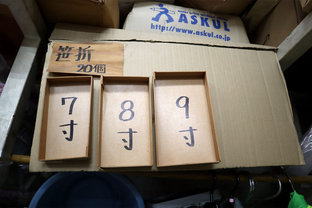 「笹折 20個」と書かれた表示の下に、7寸・8寸・9寸のサイズが書かれた3つの長方形の紙製または竹製のトレイ（折箱）が並べられている。背景には段ボール箱や「ASKUL」のロゴが入った紙袋が積まれている。包装資材を扱う店舗内の一角と思われる。