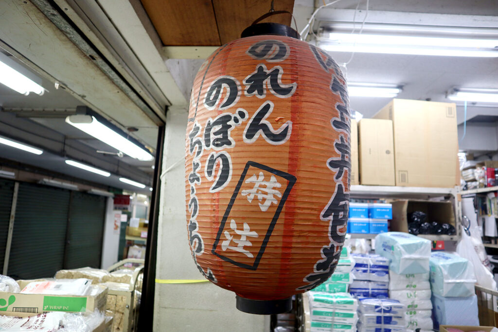 店舗の天井から吊るされた赤い提灯に、「のれん」「のぼり」「ちょうちん」「特注」などの文字が書かれており、特注対応を宣伝している。背景には段ボール箱や商品が積まれた棚があり、包装資材を取り扱う店舗の内部が見える。