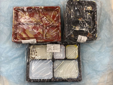 青いシートの上に並べられた3種類の仕切り付きプラスチック製弁当容器がビニールで包装されている。容器はそれぞれ色や仕切りの数が異なり、販売用のラベルシールが貼られている。業務用の弁当容器として使用される製品。